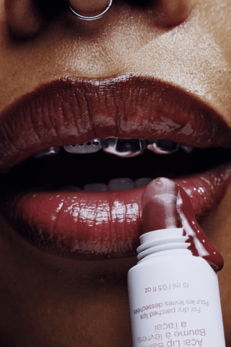 woman smiling with bleached eyebrows and tooth gems posing with topicals slick salve lip balm in açaí, makeup, skincare