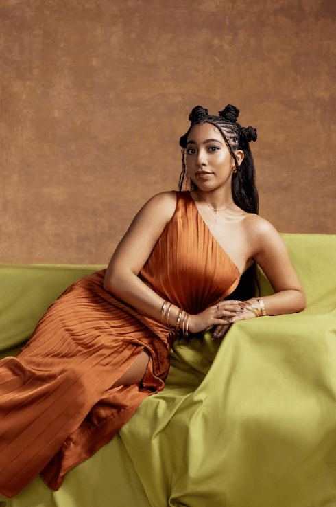 woman with braided knots on top of her head posing in orange dress, i see you, rare beauty makeup campaign