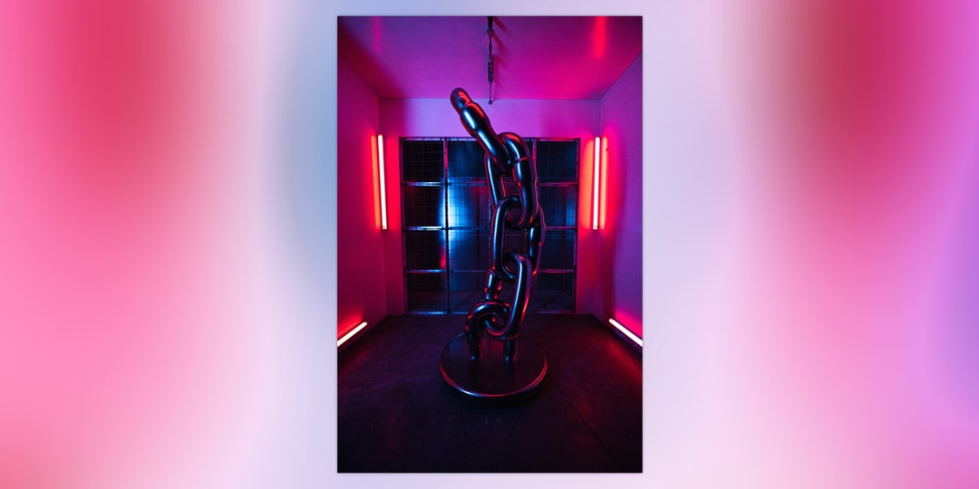 A tall chrome chain-link sculpture standing in a dark room lit by red neon tubes