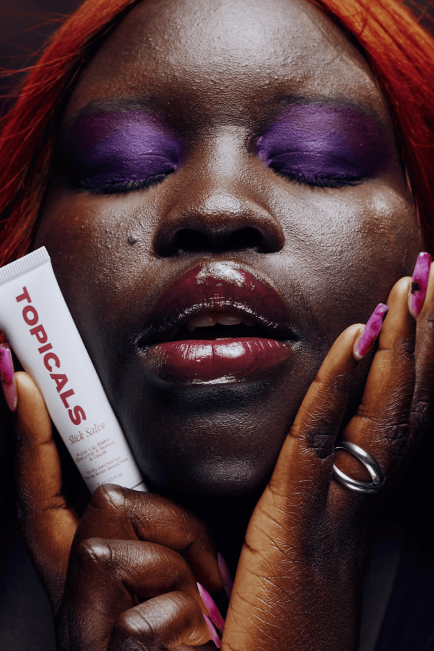 woman smiling with bleached eyebrows and tooth gems posing with topicals slick salve lip balm in açaí, makeup, skincare