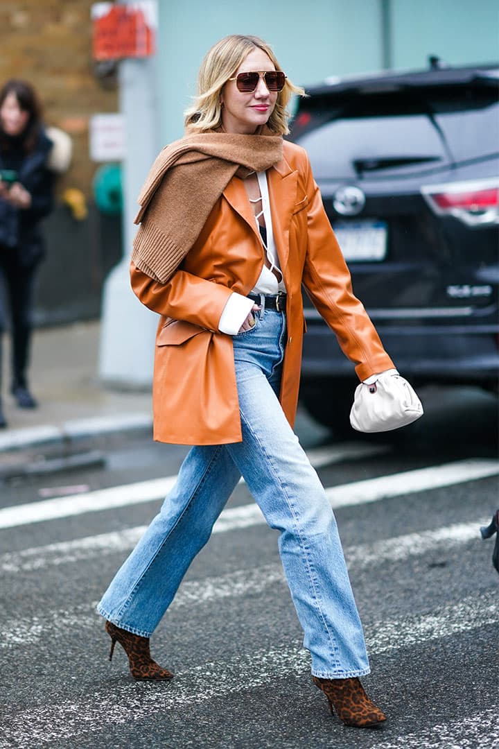 Lisa Aiken wears sunglasses, a brown wool scarf, an orange/brown leather jacket, a white top, a belt, blue denim flared pants, brown shoes, during New York Fashion Week Fall Winter 2020, on February 11, 2020 in New York City.