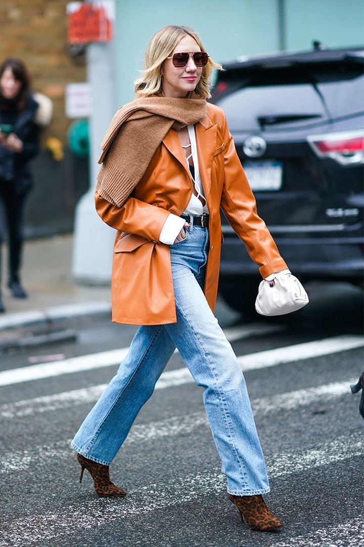 Lisa Aiken wears sunglasses, a brown wool scarf, an orange/brown leather jacket, a white top, a belt, blue denim flared pants, brown shoes, during New York Fashion Week Fall Winter 2020, on February 11, 2020 in New York City.