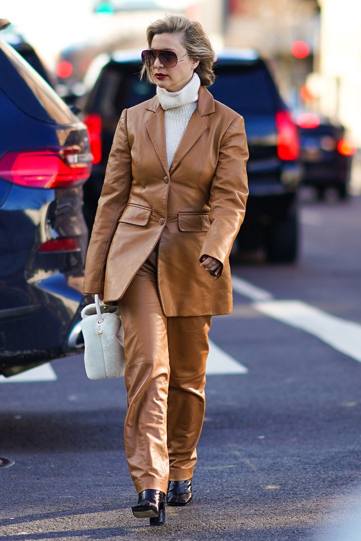 A guest wears a white wool turtleneck pullover, a brown leather jacket, leather pants, a white fluffy bag, sunglasses, outside Self Portrait, during New York Fashion Week Fall-Winter 2020