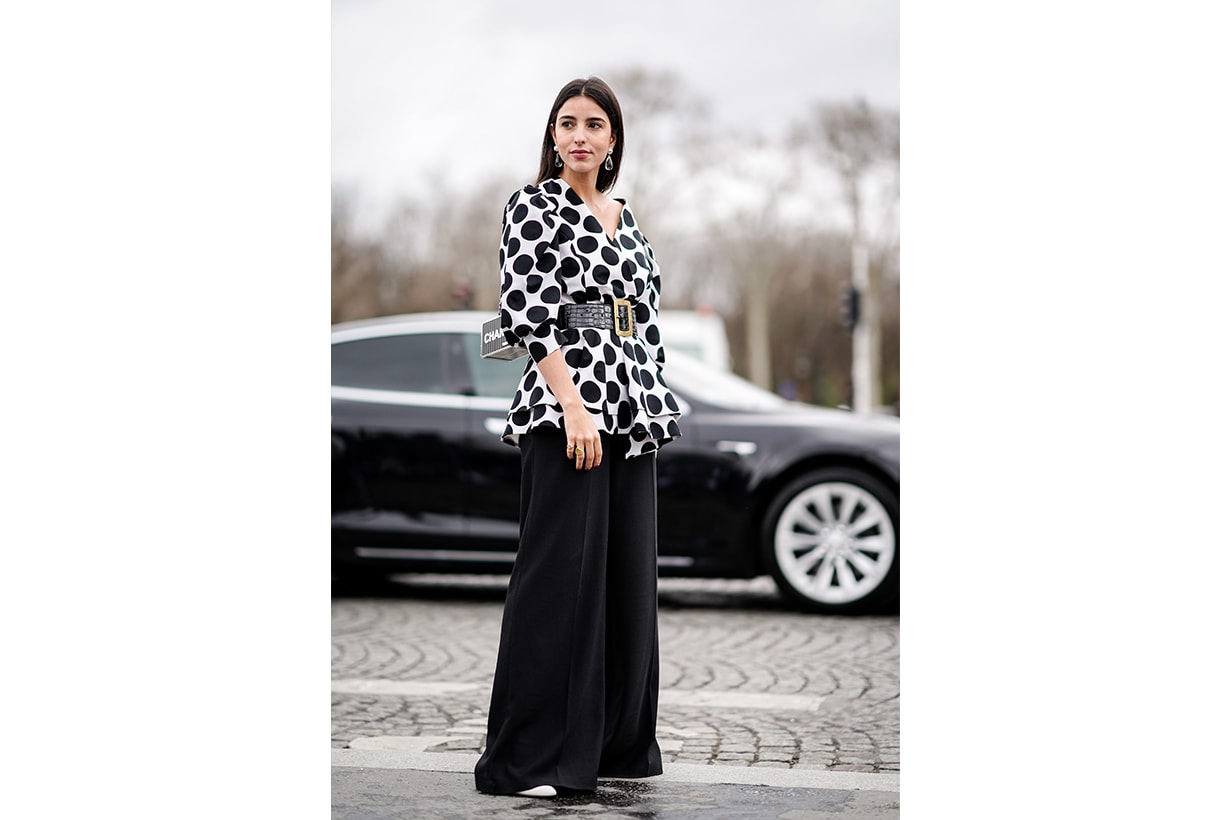 Bettina Looney wears crystal earrings, a white V-neck top with large black dots and ruffled bottom edges, a large black crocodile pattern belt with a brass buckle, black wide-leg pants, a grey metallic color Chanel box bag , outside Chanel, during Paris Fashion Week Womenswear Fall/Winter 2019/2020, on March 05, 2019 in Paris, France. 