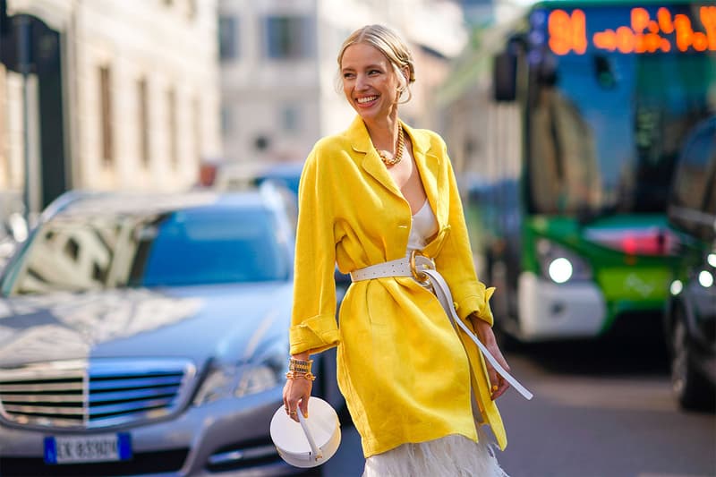 Leonie Hanne wears earrings, rings, bracelets, sunglasses, a chain necklace, a white dress with a wide neckline and decorated with white feathers, a lemon-yellow long jacket, a white belt with a gold buckle, a white Jacquemus round box bag, outside Ermanno Scervino, during Milan Fashion Week Fall/Winter 2020-2021 on February 22, 2020 in Milan, Italy.