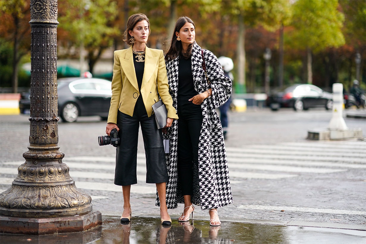 Sylvia Haghjoo (L) wears Chanel earrings, a Chanel necklace, a yellow jacket, a black top, black leather wide-legs crop pants, a grey clutch, Chanel slingback pumps, holds a Canon camera with a 24-70mm zoom lens ; Julia Haghjoo (R) wears Chanel earrings, a bracelet, a black and white houndstooth oversized coat, a black top, black crop pants, white heeled sandals, outside Chanel, during Paris Fashion Week - Womenswear Spring Summer 2020, on October 01, 2019 in Paris, France.