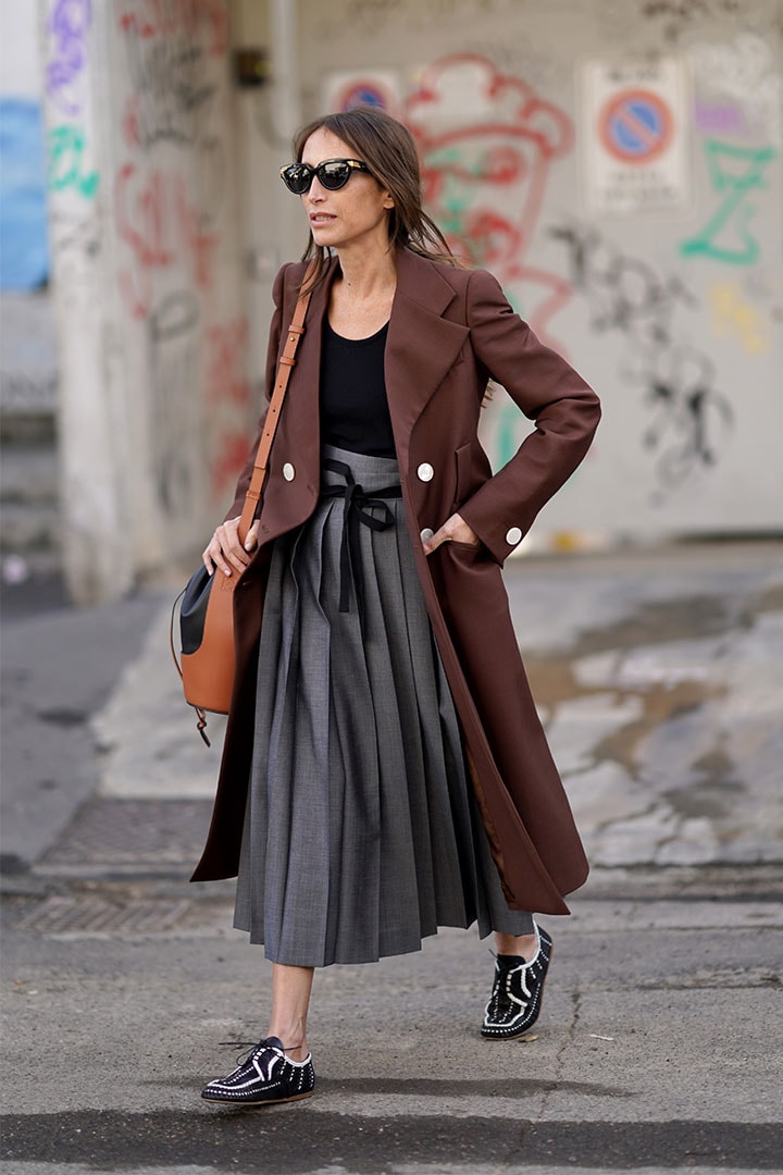 Chloe Harrouche wears sunglasses, a brown long jacket, a black top, a gray pleated skirt, a leather brown and black Loewe bag, black and white shoes, outside Marni, during Milan Fashion Week Fall/Winter 2020-2021 on February 21, 2020 in Milan, Italy.