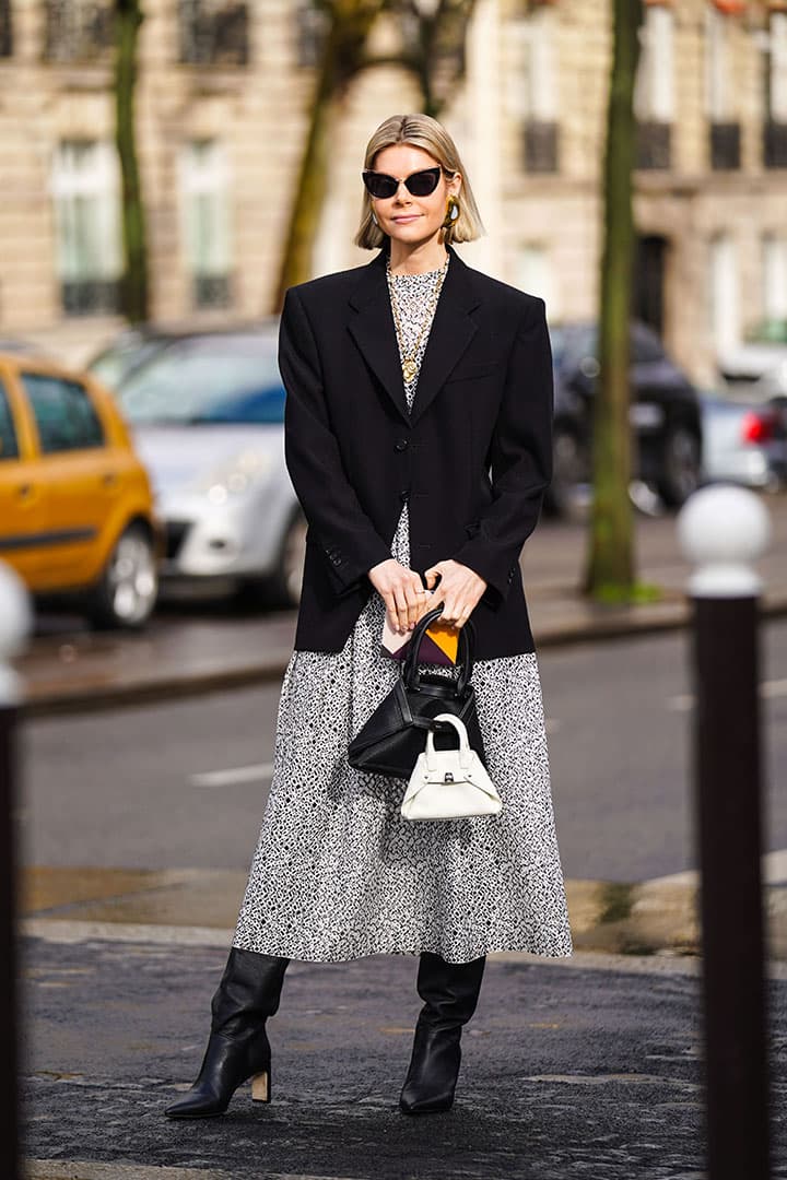 Kate Tik wears sunglasses, earrings, necklaces, a white and black mesh dress, a black jacket, a black bag and a white one, black pointy heeled boots, outside Akris, during Paris Fashion Week - Womenswear Fall/Winter 2020/2021, on March 02, 2020 in Paris, France.